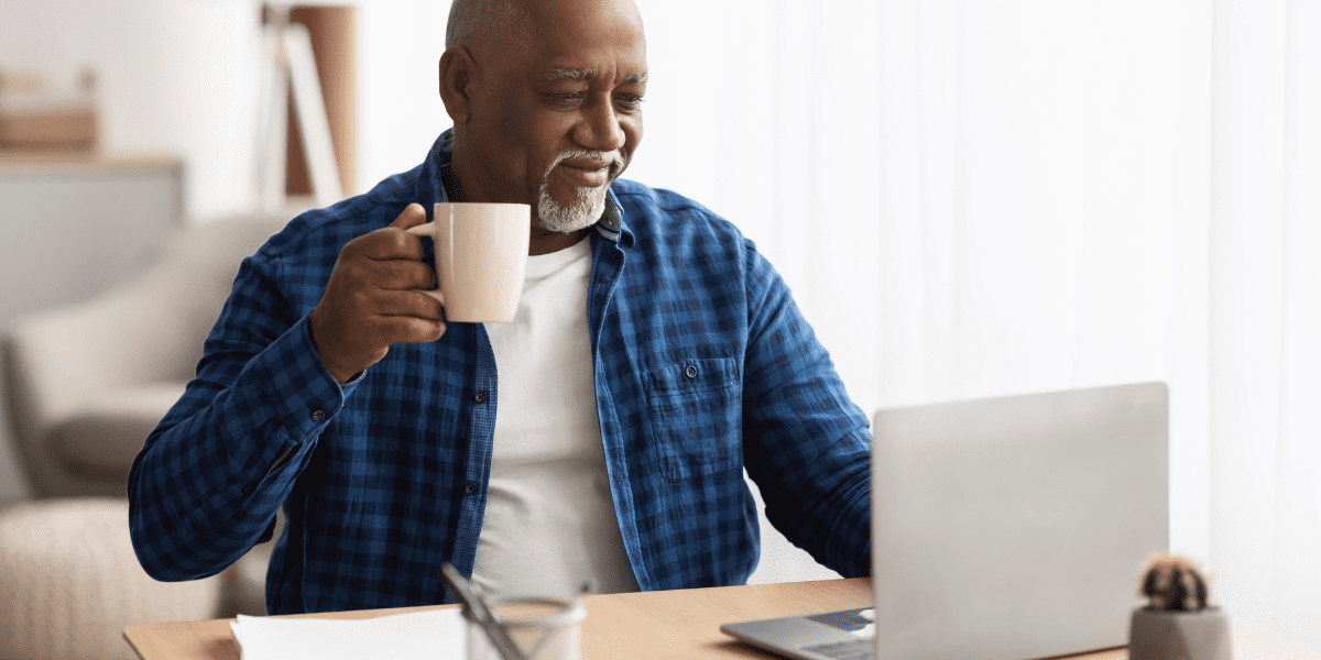 Retired Man Holding Mug and Smiling While Looking At A Laptop In His Living Room