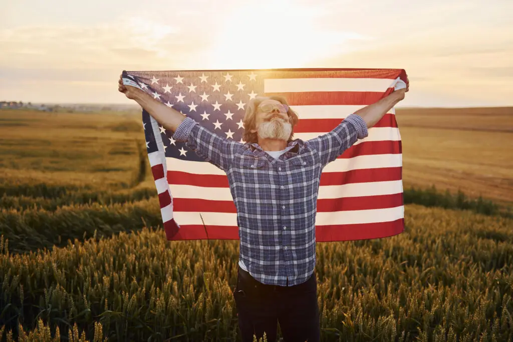 Patriotic senior man wearing glasses and smiling while holding American Flag in a field of wheat